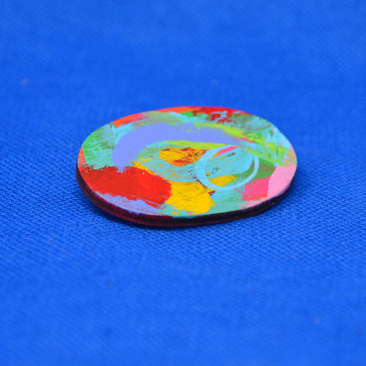 An oval-shaped painted wooden brooch featuring an abstract multicolor design with red, orange, yellow, green, blue, and purple brushstrokes, displayed on a textured blue fabric background.