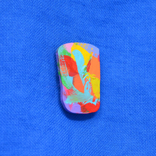 An irregular shaped wooden brooch featuring a colorful bold brushstrokes abstract paint design with pink, blue, orange, purple, green and red brushstrokes, set against a textured blue background.