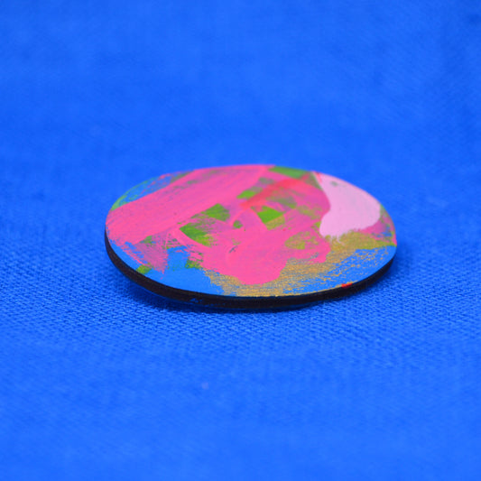 An oval, hand-painted wooden brooch featuring a vibrant multicolor abstract pattern of pink, gold, blue, and green brushstrokes, set against a textured bright blue background.
