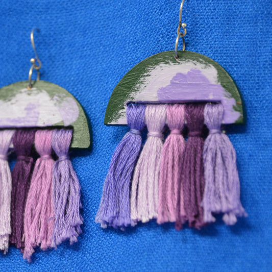 A pair of handmade dangle earrings with semi-circular wooden tops painted in green and lavender, hanging from silver fishhook wires. Each top is adorned with five tassels in various shades of purple and lavender, arranged against a bright blue textured fabric background.