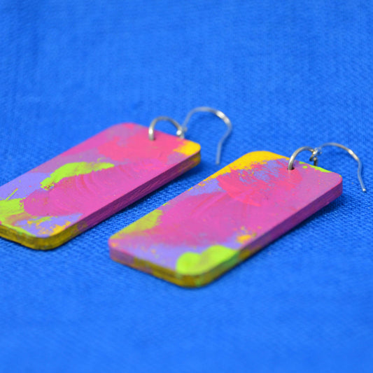 A pair of rectangular drop earrings with a vibrant pink, yellow, and green abstract paint design, attached to silver fishhook wires, displayed on a textured blue background.