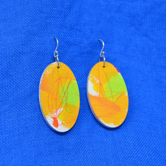 A pair of oval drop earrings with abstract painted designs in orange, yellow, green, and white, suspended from silver fishhook wires, displayed on a textured blue background.