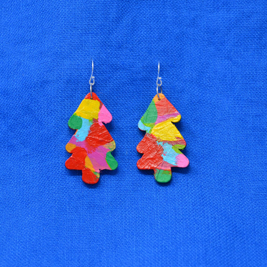 A pair of tree-shaped drop earrings made from colorful, textured paper pieces in shades of red, orange, yellow, green, pink, and blue, hanging from silver fishhook wires against a bright blue textured fabric background.