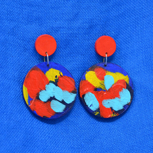 A pair of handmade drop earrings with bright red circular studs and circle dangles featuring a blue, red and yellow abstract painted design, displayed on a textured blue background.