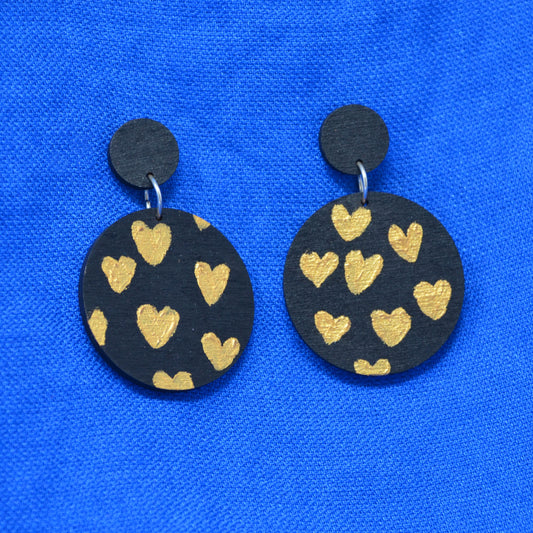 A pair of handmade drop earrings featuring round black wooden discs decorated with hand-painted gold hearts, displayed on a vibrant blue textured fabric background.