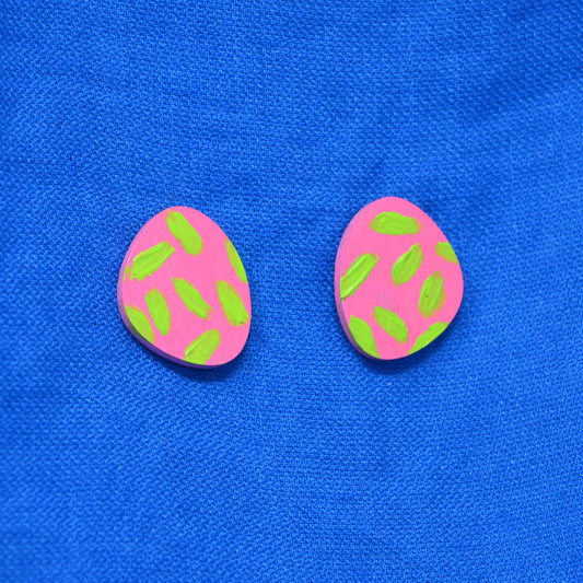 A pair of pink oval stud earrings with green painted detail, displayed on a textured blue fabric background.
