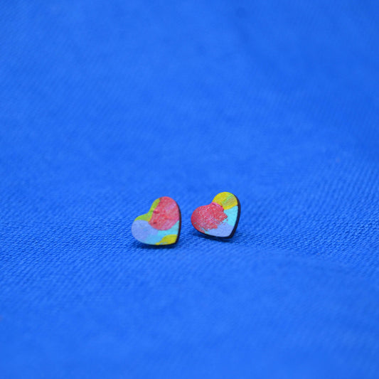 A pair of small heart-shaped stud earrings with a colorful, textured paint-splatter design in pink, yellow, blue, and green, resting on a textured blue background.