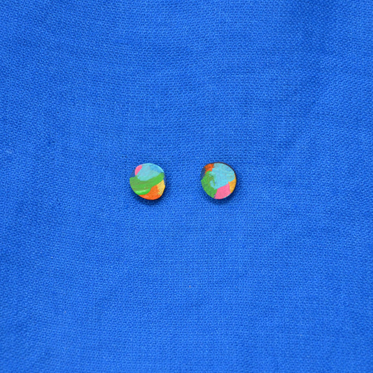 A pair of small oval stud earrings with a colorful abstract marbled pattern in green, blue, pink, and orange, displayed on a textured blue fabric background.