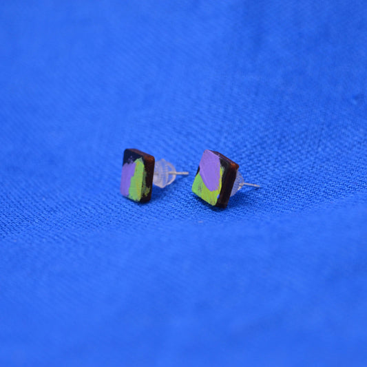 A pair of wooden square stud earrings with a black base and an abstract paint design in purple and green, displayed on a textured blue background.