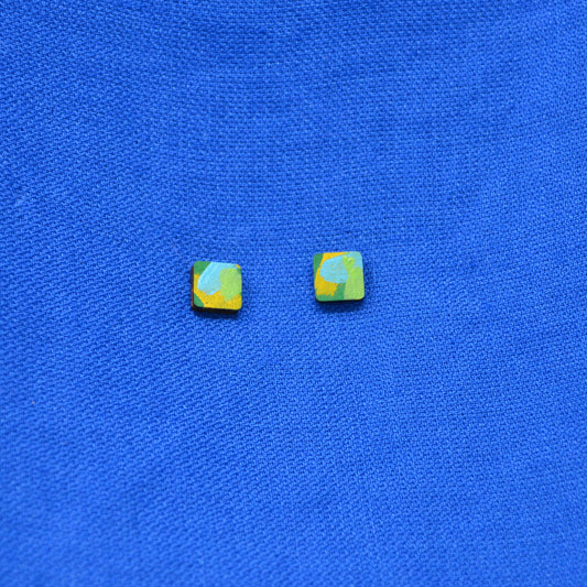 A pair of small square stud earrings with a marbled pattern of yellow, green, and light blue, displayed on a textured blue fabric background.