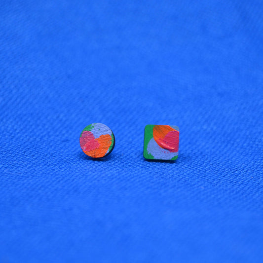 A pair of small stud earrings, one circular and one square, featuring a colorful abstract paint pattern in orange, red, green, and blue, displayed on a textured blue background.