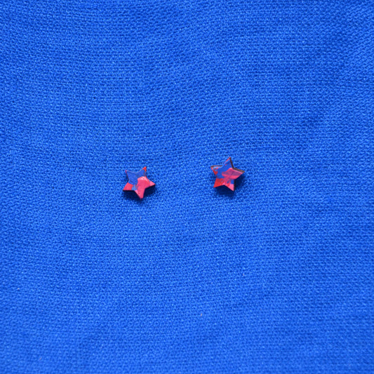Two small wooden star-shaped stud earrings with a pink and blue painted pattern, displayed on a textured blue background.
