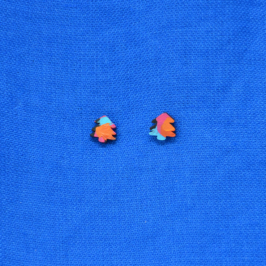 A pair of small multicolor stud earrings shaped like stylized trees, featuring orange, pink, and light blue with black outlines, placed on a textured blue fabric background.