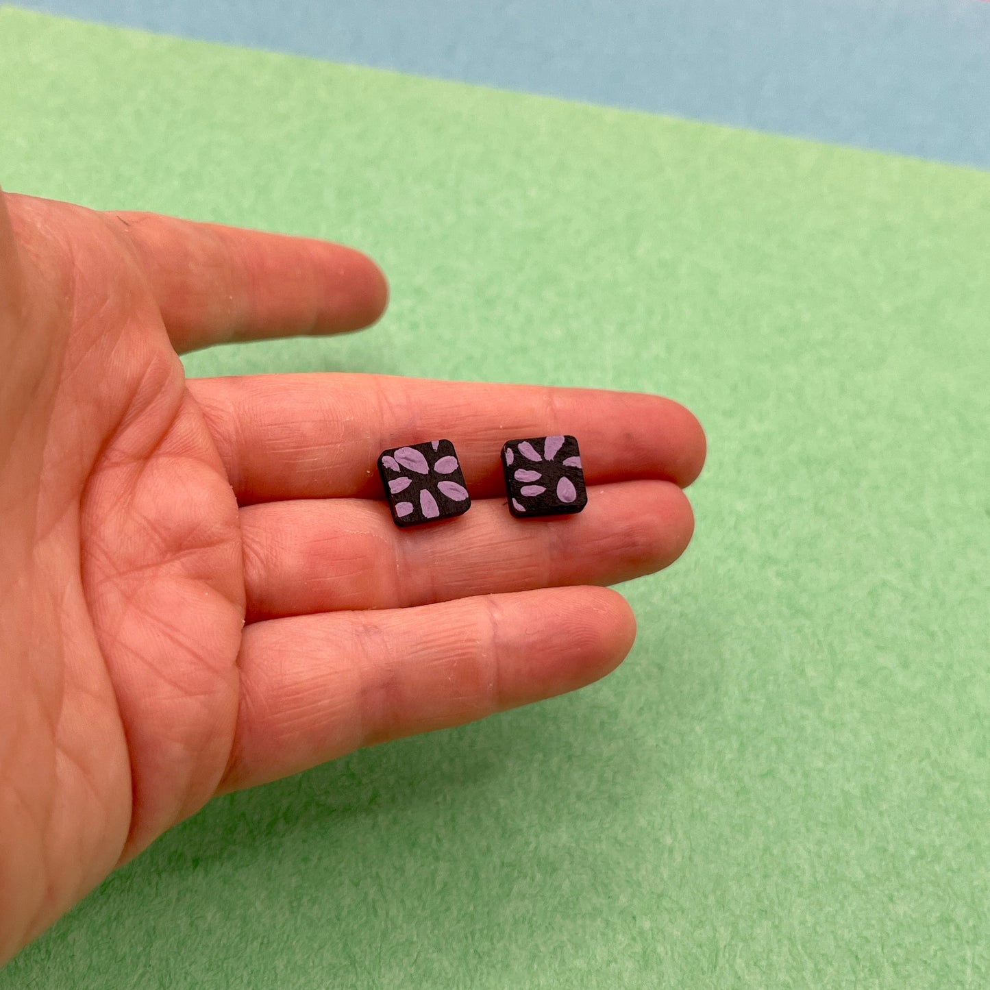 Two small black earrings with purple patterns held in a hand against a green background