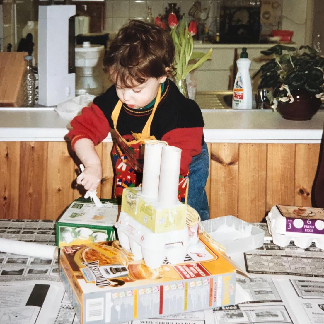 A young child wearing a red and black shirt and an orange apron stands at a kitchen counter, surrounded by household items, a newspaper-covered table, and a box of cereal in a retro-style kitchen scene.