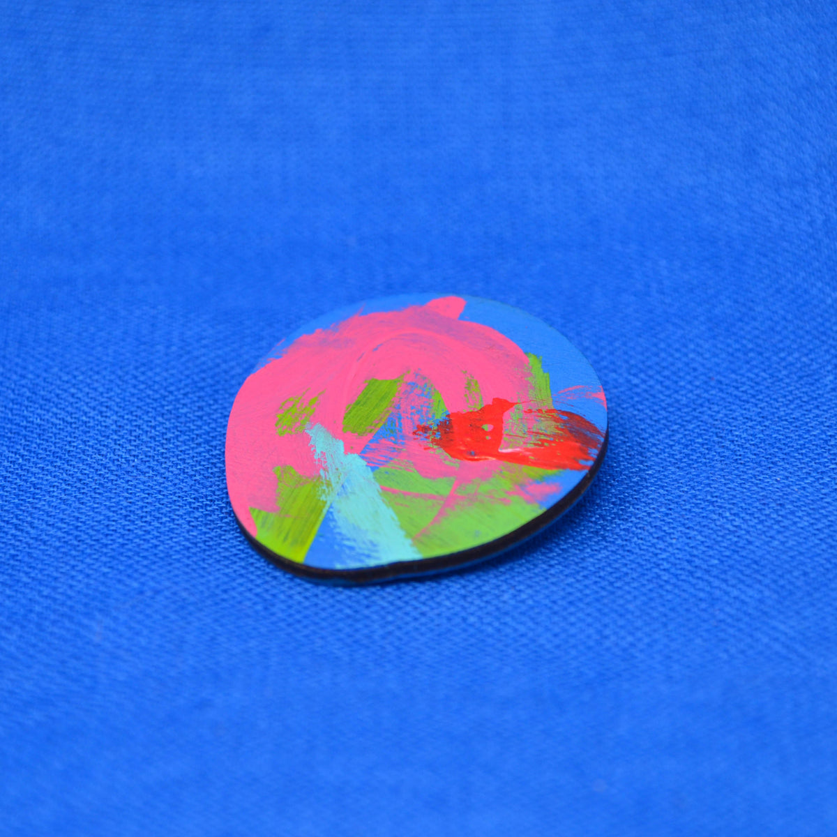 An oval pin or badge with a colorful abstract paint design, featuring pink, blue, green, and red brushstrokes on a wooden base, set against a textured blue background.