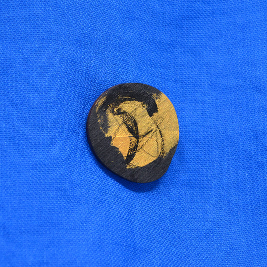 An round-shaped painted wooden brooch featuring an abstract multicolor design with black and gold brushstrokes, displayed on a textured blue fabric background.
