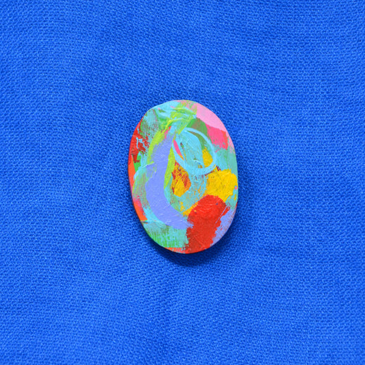 An oval-shaped painted wooden brooch featuring an abstract multicolor design with red, orange, yellow, green, blue, and purple brushstrokes, displayed on a textured blue fabric background.
