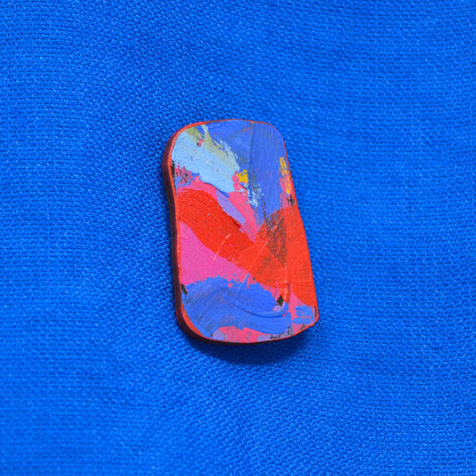 An irregular shaped wooden brooch featuring a colorful bold brushstrokes abstract paint design with pink, blue and red brushstrokes, set against a textured blue background.