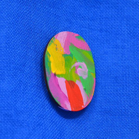 An oval  wooden brooch featuring a colorful abstract paint design with pink, green, yellow and red brushstrokes, set against a textured blue background.