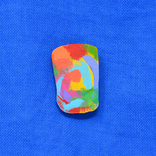 An irregular rectangle hand-painted wooden brooch featuring a vibrant multicolor abstract pattern of orange, red, green, yellow, blue and purple brushstrokes, set against a textured bright blue background.