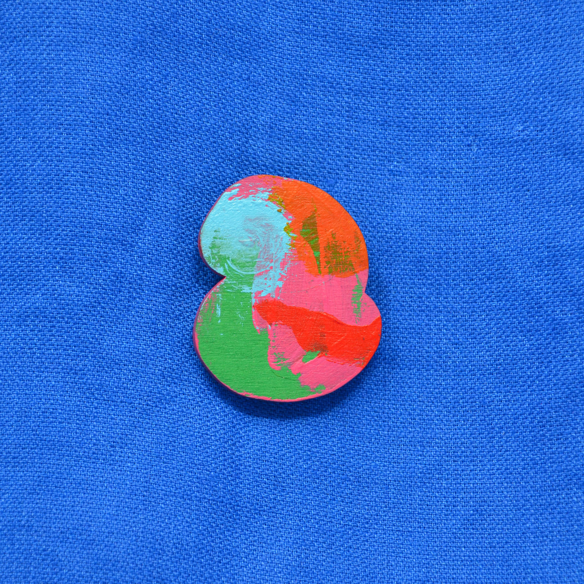 A small, irregularly shaped button or badge with a colorful abstract paint pattern in red, green, and light blue, set against a textured blue fabric background.