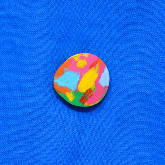 A round, hand-painted wooden brooch featuring a vibrant multicolor abstract design, displayed on a textured blue fabric background.