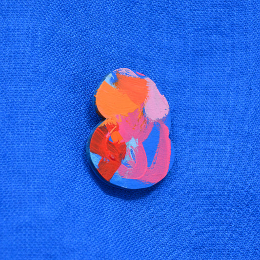 A pin or badge with abstract multicolor paint strokes in orange, pink, red, and blue, placed on a bright blue textured fabric background.