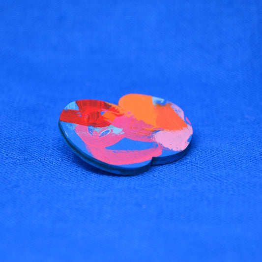 A pin or badge with abstract multicolor paint strokes in orange, pink, red, and blue, placed on a bright blue textured fabric background.