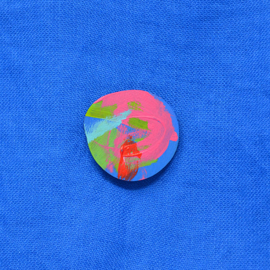 An round-shaped painted wooden brooch featuring an abstract multicolor design with blue, pink, green and red brushstrokes, displayed on a textured blue fabric background.