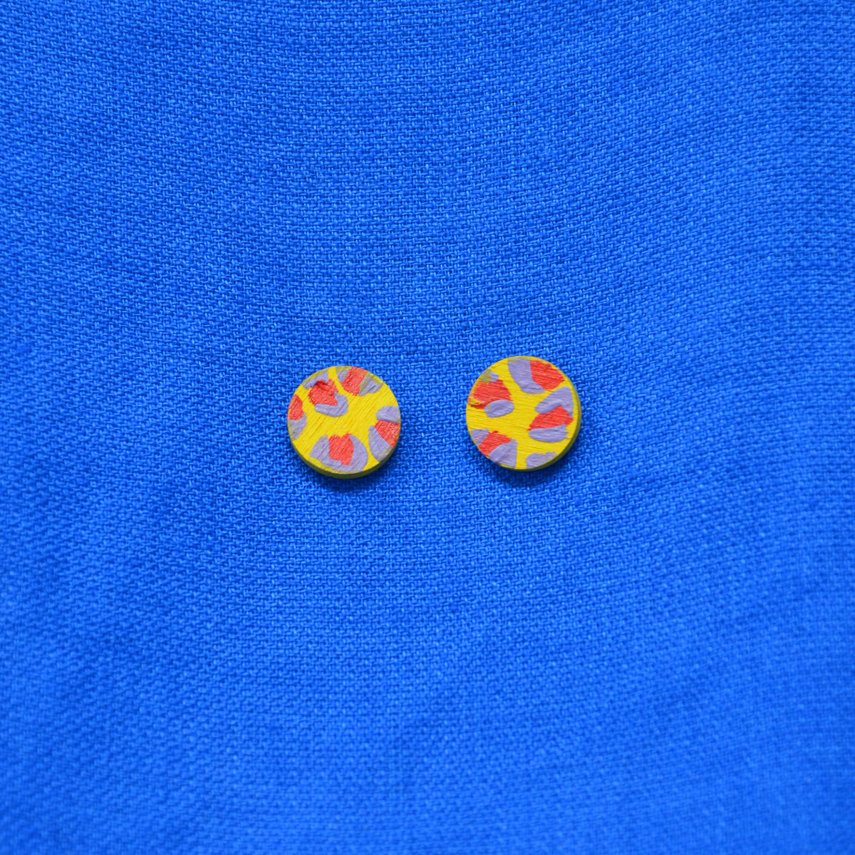 A pair of round yellow earrings with a red and muted purple abstract leopard print pattern, displayed on a textured blue fabric background.