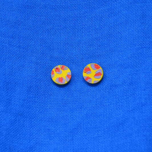 A pair of round yellow earrings with a red and muted purple abstract leopard print pattern, displayed on a textured blue fabric background.