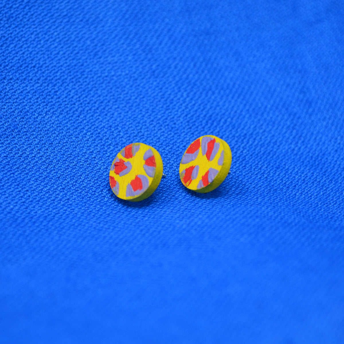 A pair of round yellow earrings with a red and muted purple abstract leopard print pattern, displayed on a textured blue fabric background.