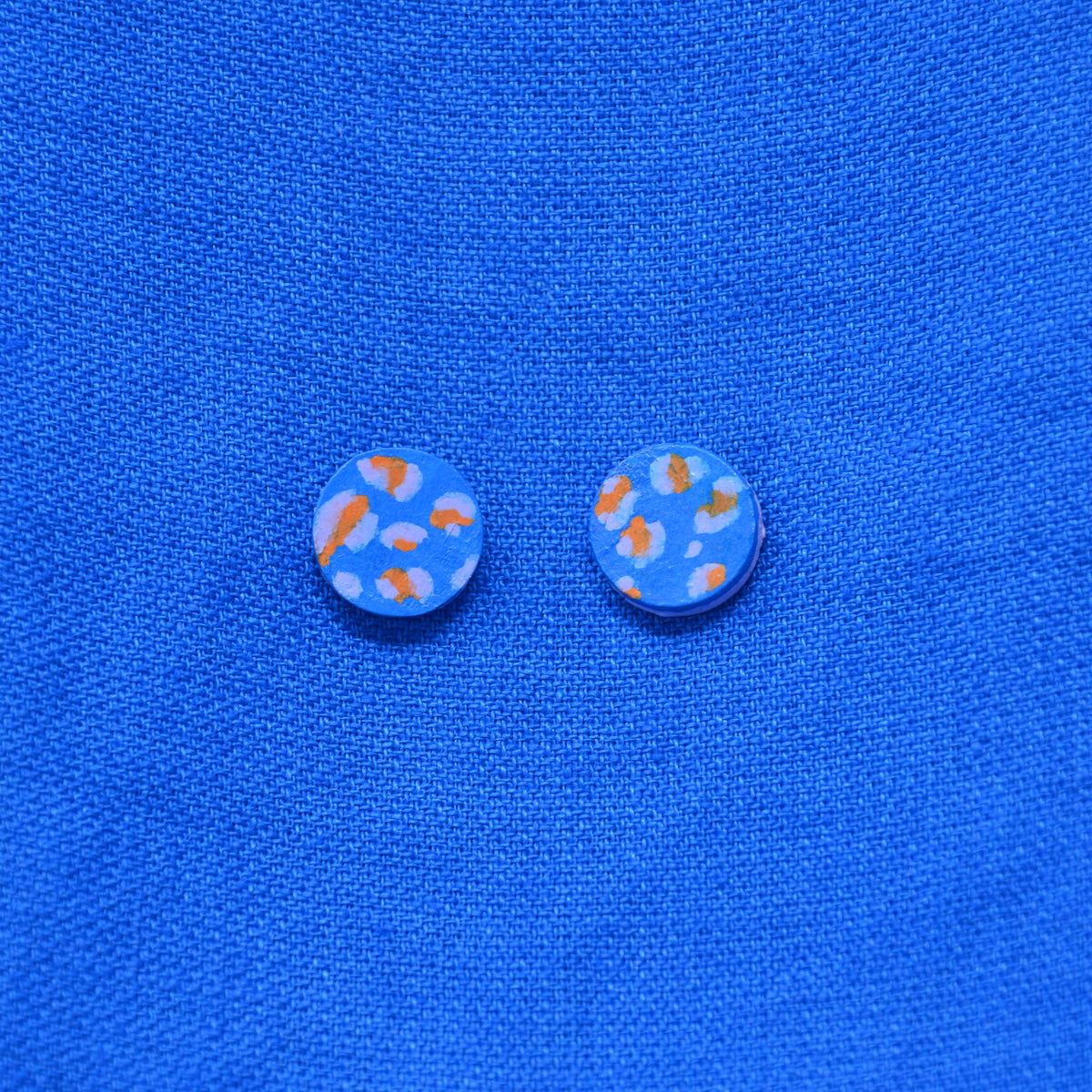 A pair of round stud earrings with a light blue background and orange and pale blue abstract spots, displayed on a textured blue fabric background.