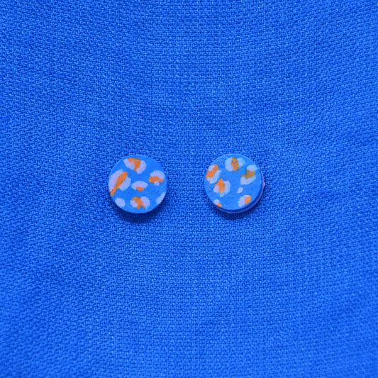 A pair of round stud earrings with a light blue background and orange and pale blue abstract spots, displayed on a textured blue fabric background.