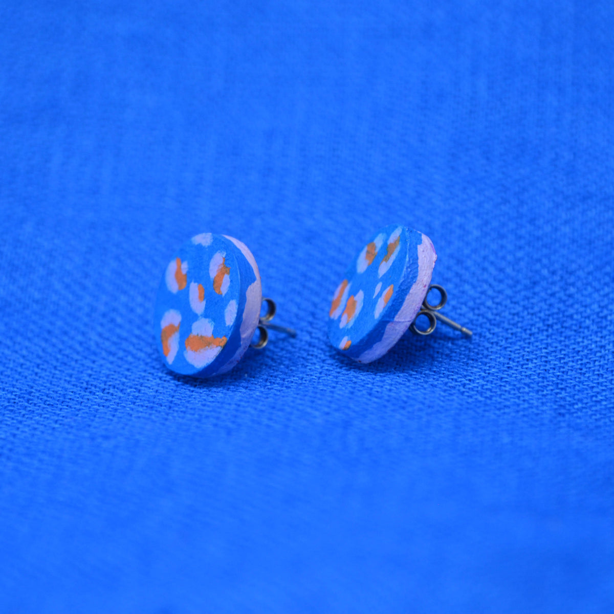 A pair of wooden round stud earrings with a light blue background and orange and pale blue abstract spots, displayed on a textured blue fabric background.
