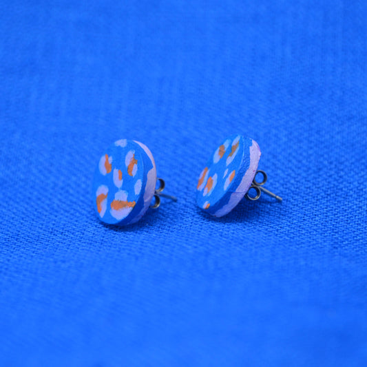 A pair of wooden round stud earrings with a light blue background and orange and pale blue abstract spots, displayed on a textured blue fabric background.