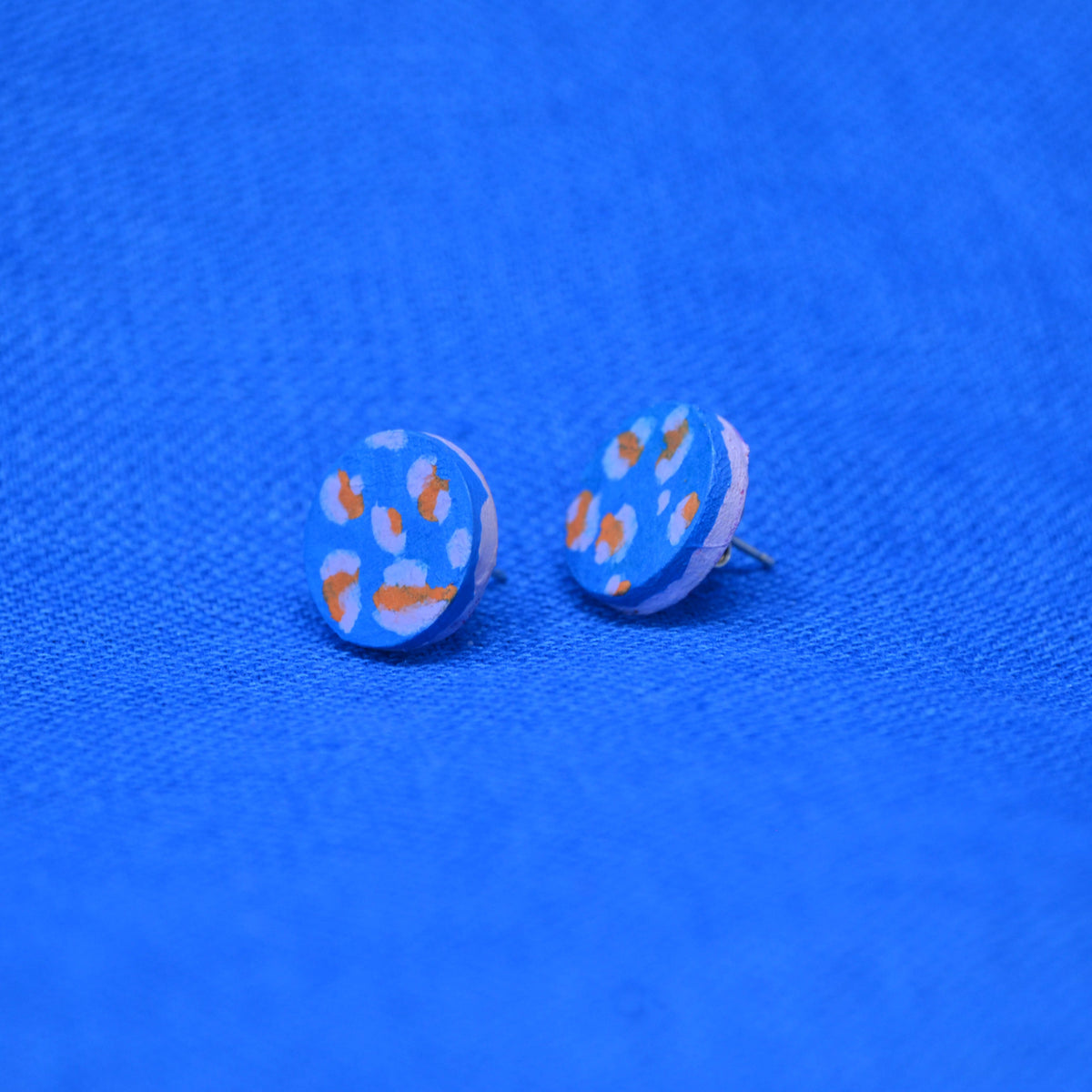 A pair of round stud earrings with a light blue background and orange and pale blue abstract leopard like spots, displayed on a textured blue fabric background.