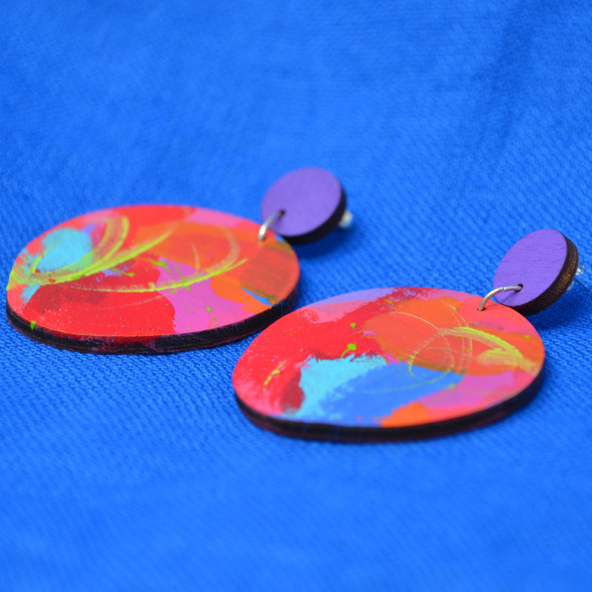 A pair of colorful wooden dangle earrings with abstract painted designs in red, orange, pink, blue, and yellow, featuring purple circular studs, displayed on a textured blue background.