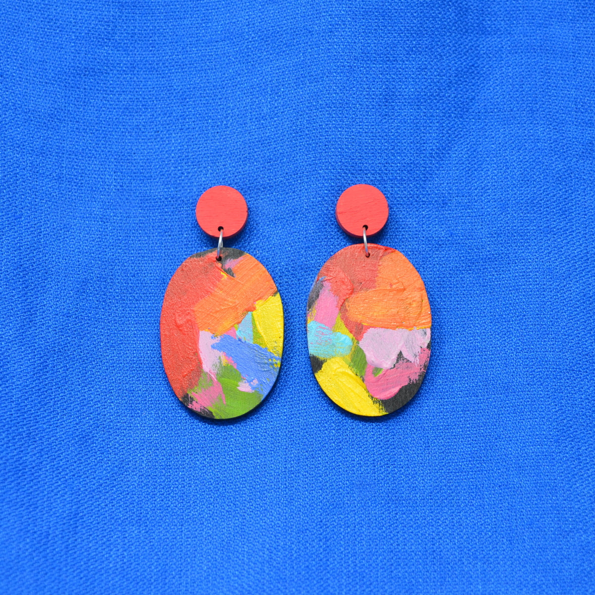 A pair of drop earrings with bright coral-red circular studs and oval dangles featuring an abstract multicolor paint-stroke pattern, displayed on a textured blue background.