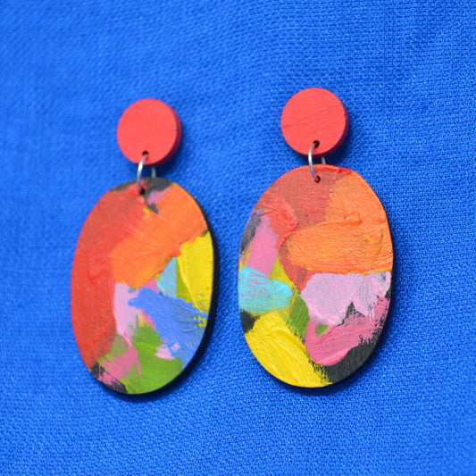 A pair of handmade oval drop earrings with bright red circular studs and multicolored abstract painted wooden dangles, displayed on a textured blue background.