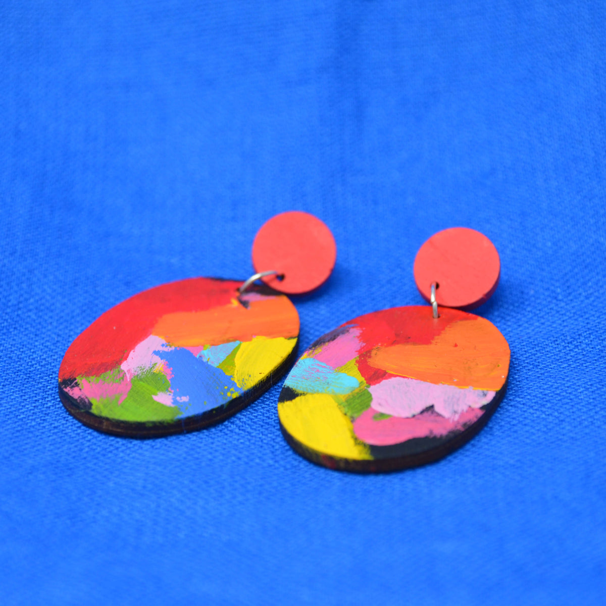 A pair of handmade dangle earrings with flat, irregular oval-shaped wooden discs painted with a colorful abstract brushstroke pattern, hanging from bright red circular studs, displayed on a textured blue background.