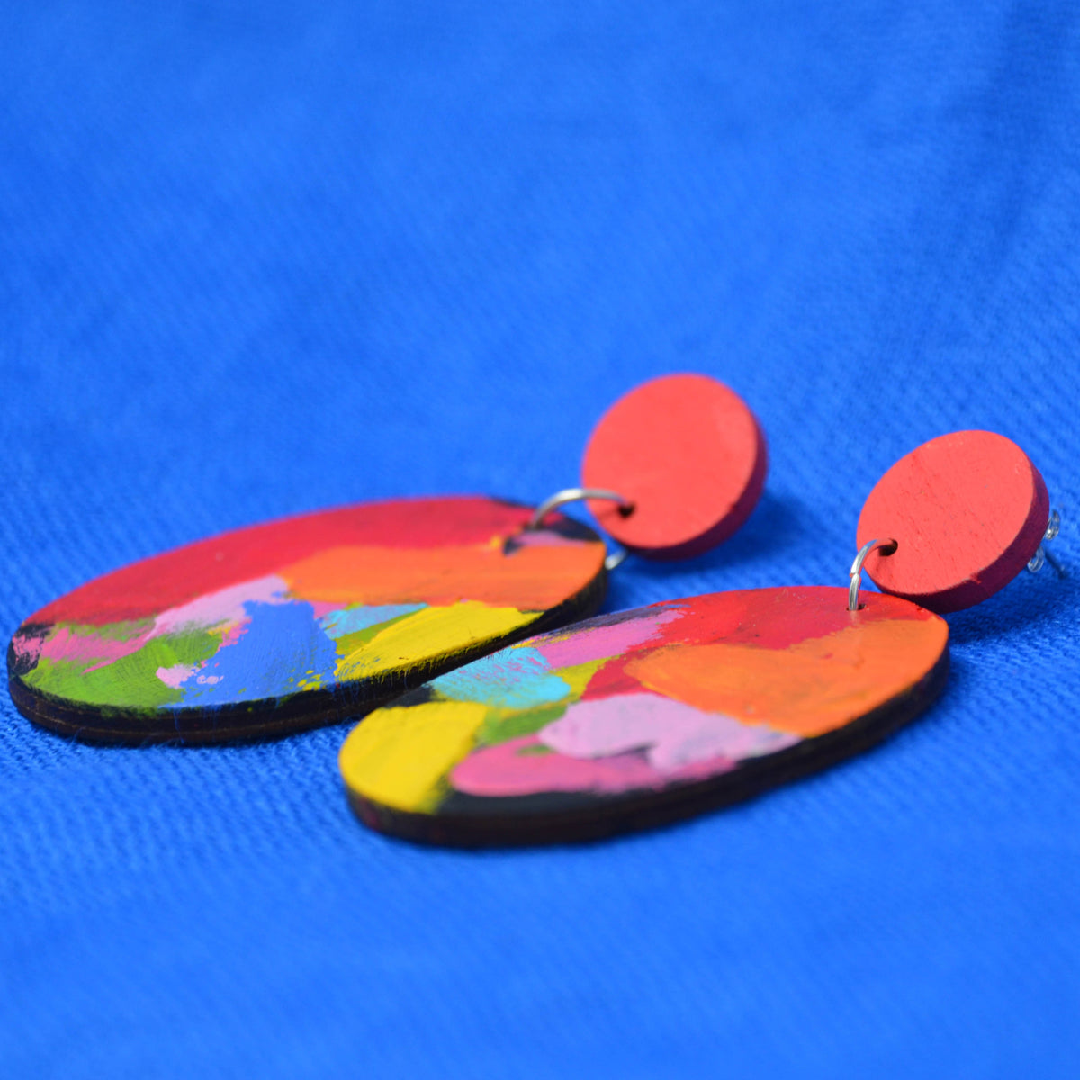 A pair of handmade wooden dangle earrings with colorful abstract painted patterns and solid red circular studs, displayed on a textured blue background.