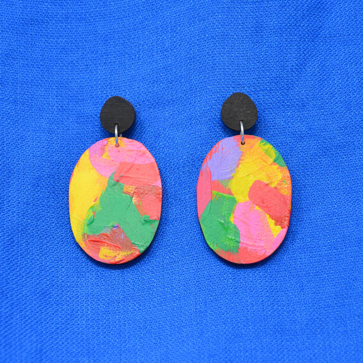 A pair of handmade oval drop earrings with colorful, textured abstract paint patterns in pink, yellow, green, and purple, suspended from black round studs against a bright blue textured background.