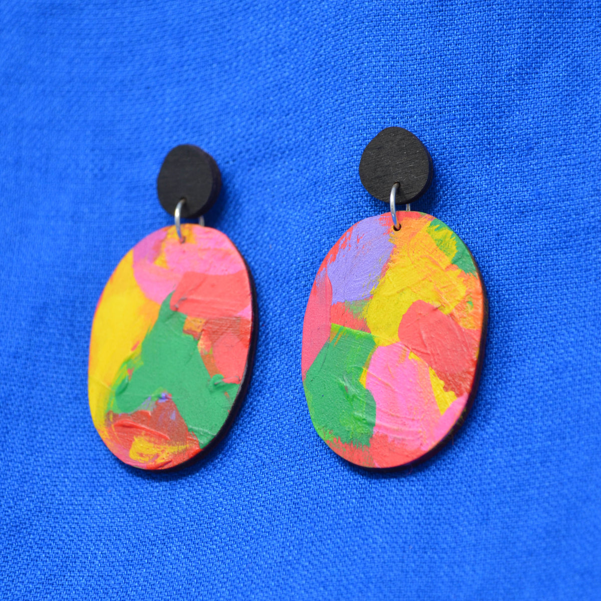 A pair of handmade oval drop earrings with colorful, textured paint-like patterns in pink, yellow, green, and purple, suspended from black circular studs against a bright blue textured fabric background.