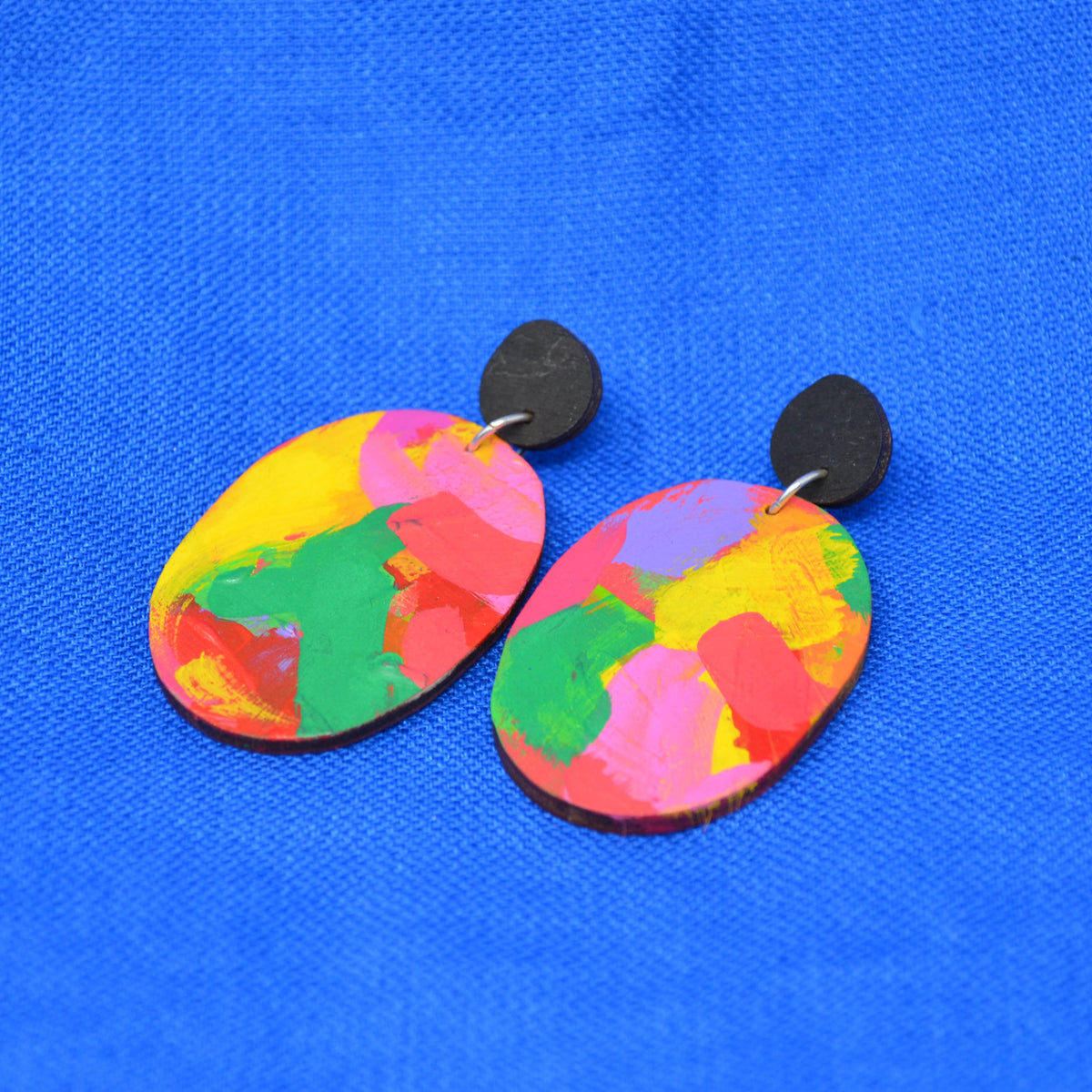 A pair of handmade oval drop earrings featuring a vibrant, multicolored abstract paint pattern in pink, yellow, green, and red, suspended from matte black teardrop-shaped studs against a bright blue textured background.