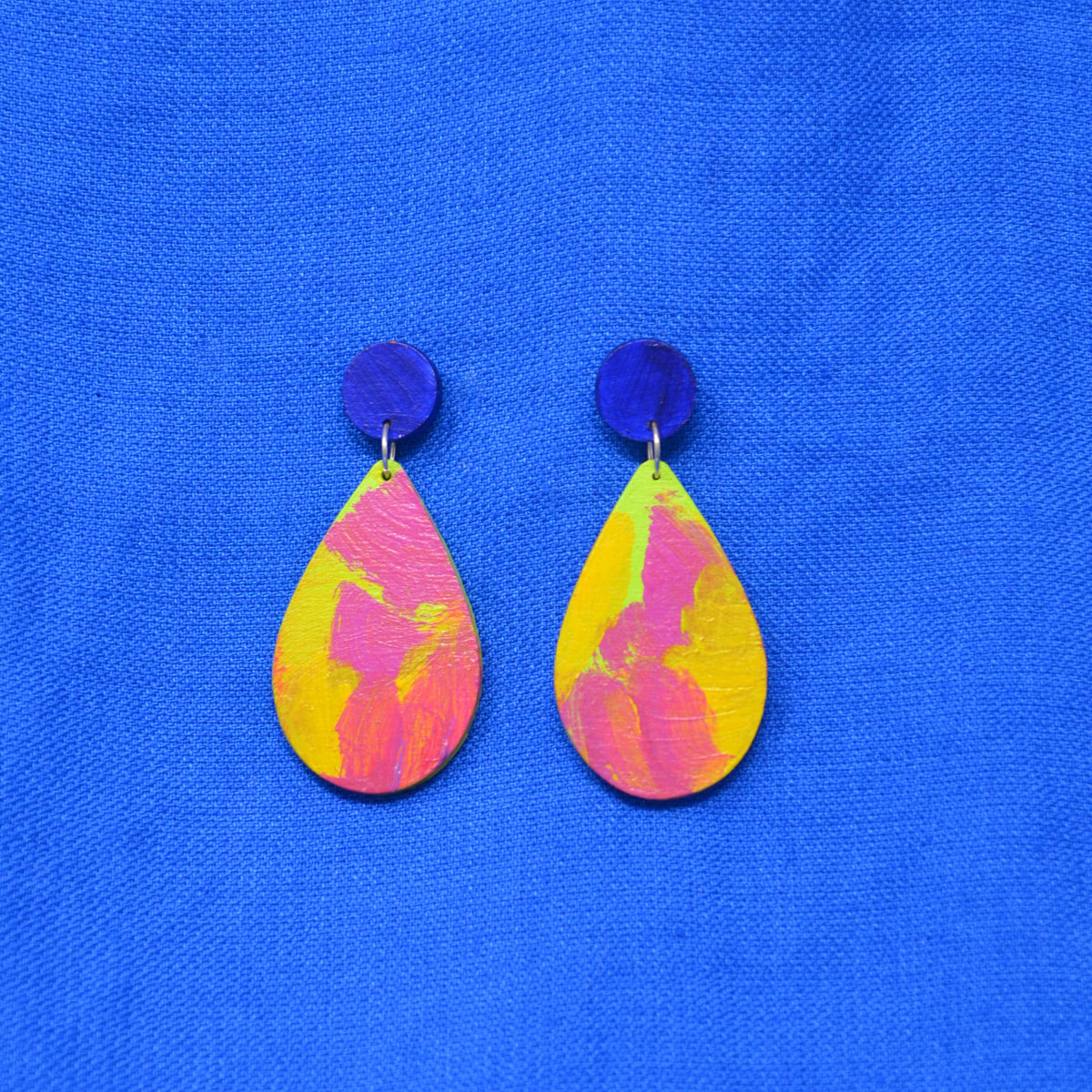 A pair of handmade drop earrings with dark blue circular studs and teardrop shaped dangles featuring a pink, green and yellow abstract painted design, displayed on a textured blue background.
