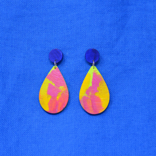A pair of handmade drop earrings with dark blue circular studs and teardrop shaped dangles featuring a pink, green and yellow abstract painted design, displayed on a textured blue background.