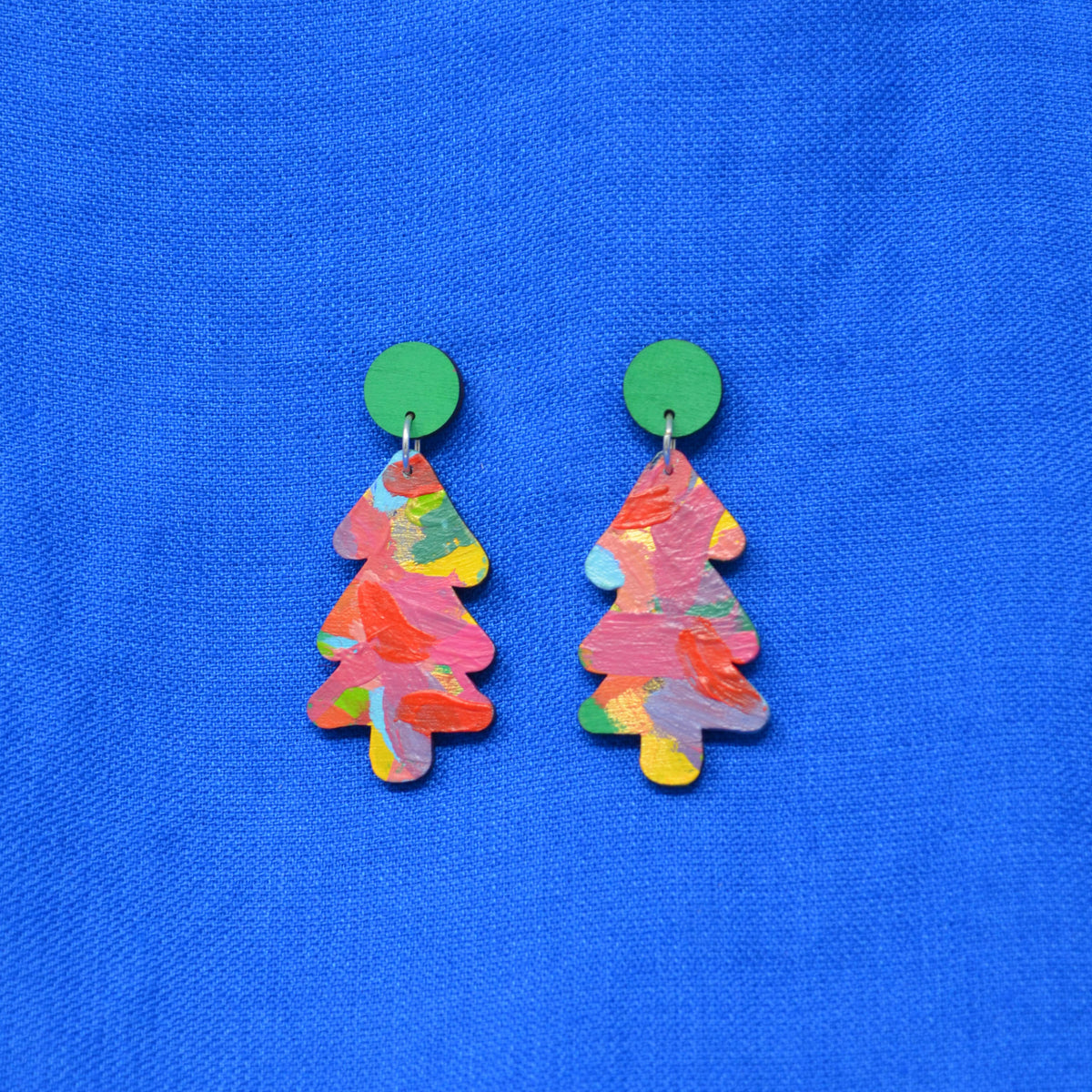 A pair of Christmas tree-shaped earrings with a colorful, textured paint-stroke pattern in pink, orange, yellow, and green, hanging from solid green circular studs against a bright blue textured fabric background.