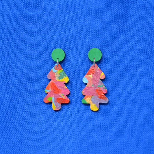 A pair of Christmas tree-shaped earrings with a colorful, textured paint-stroke pattern in pink, orange, yellow, and green, hanging from solid green circular studs against a bright blue textured fabric background.